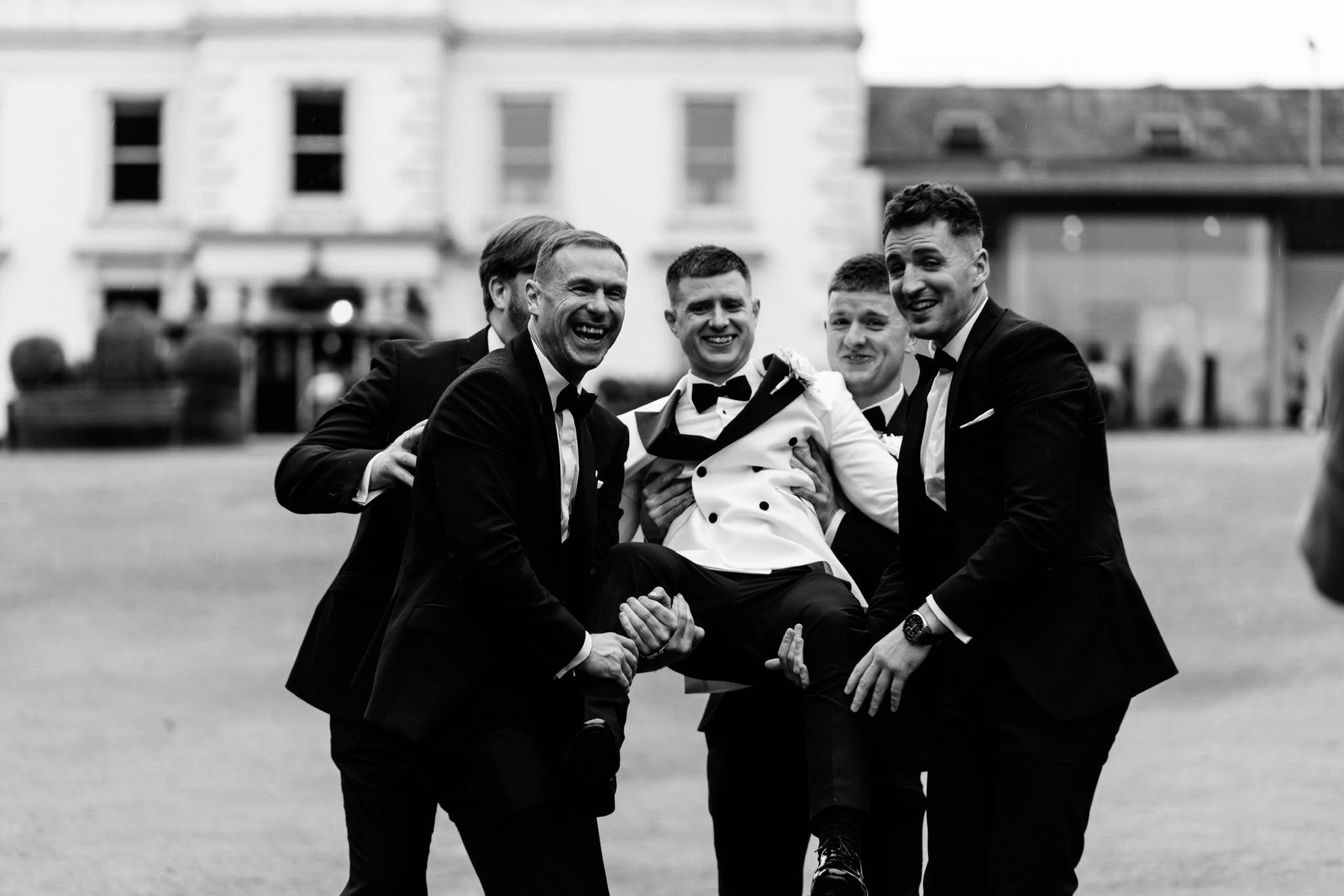 Groomsmen in black tie laughing and lifting one of the party outside the house.