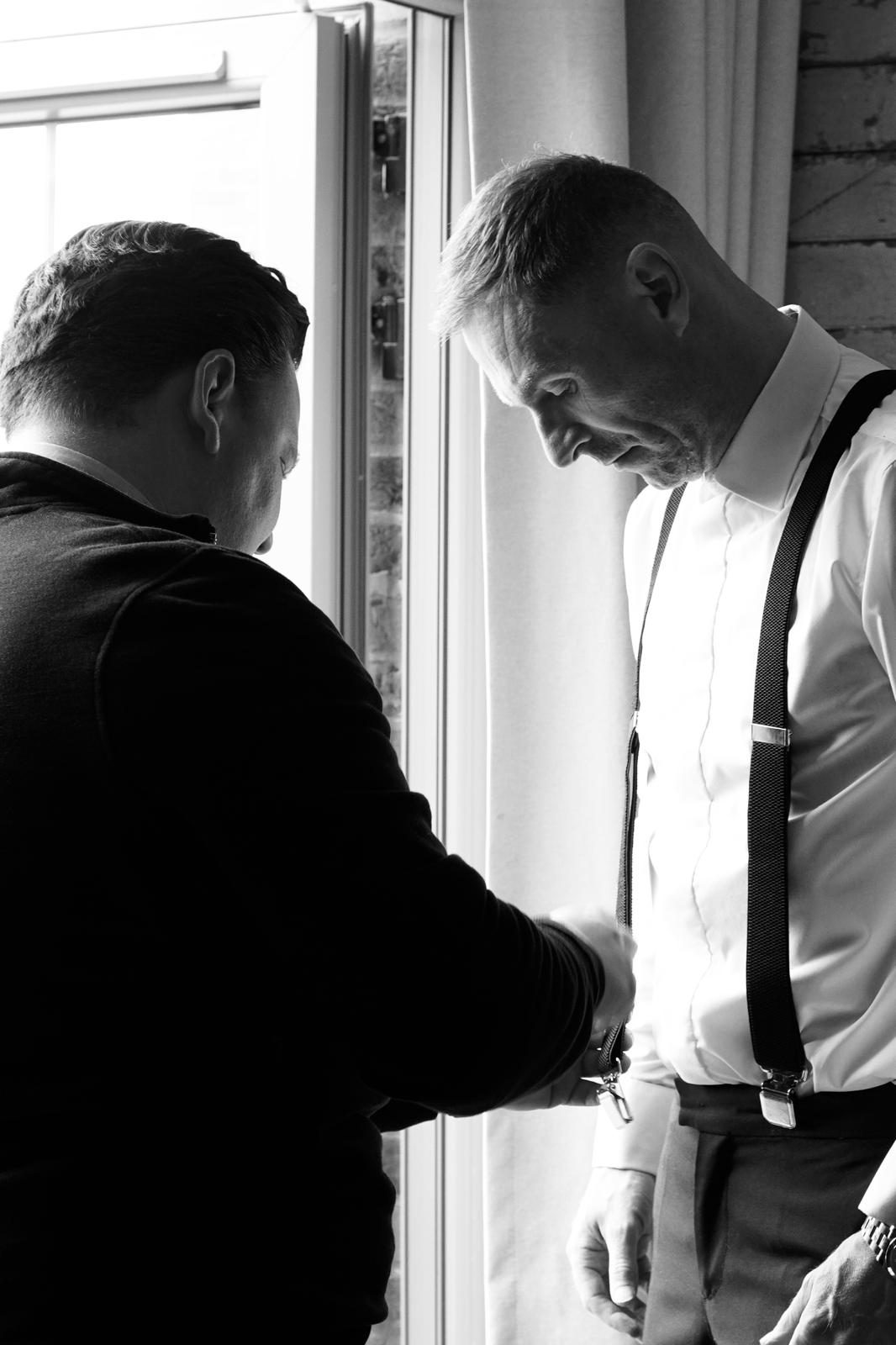 Raymond fastening a cufflink at the wrist of a groomsman in shirt and braces.