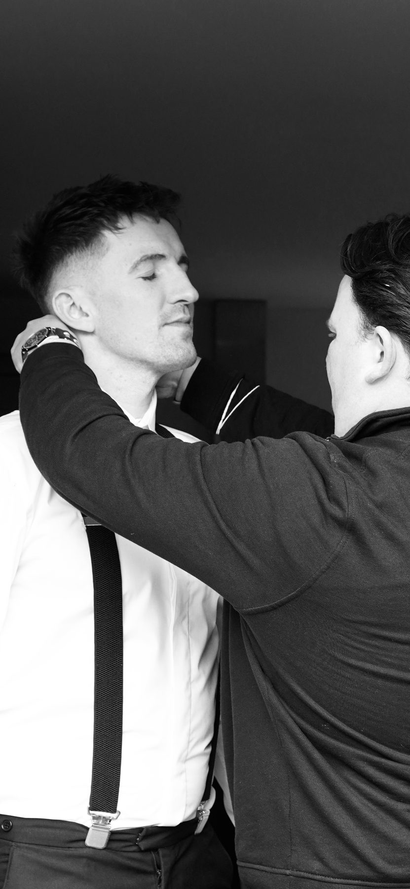 Raymond fastening a bow tie from behind for a groomsman in braces.