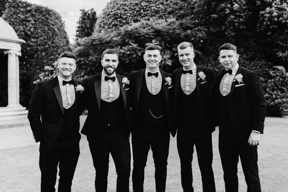 Six men in black tie lined up in front of a hedge, dressed and ready.