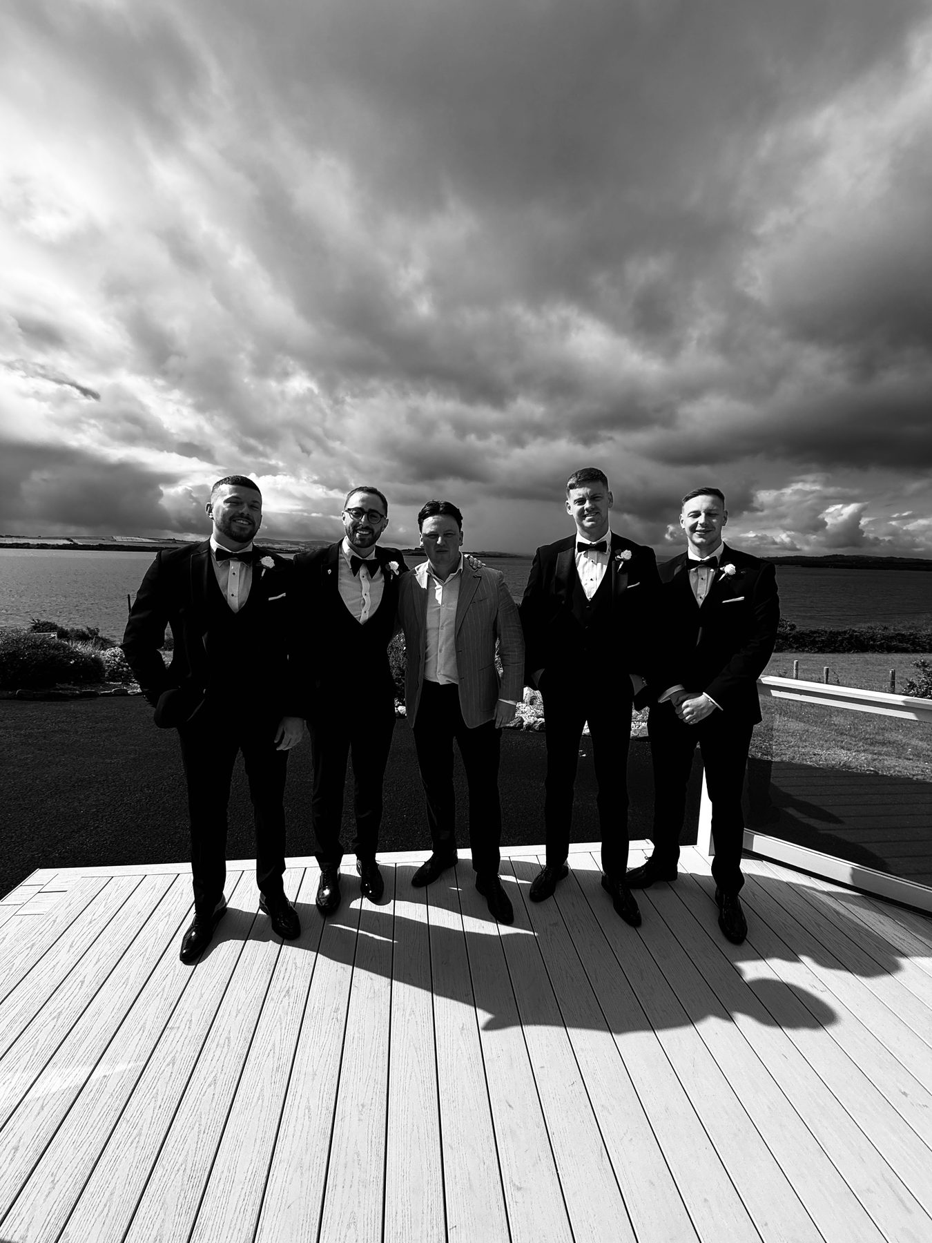 Five men in black tie on decking under a dramatic sky by the water.